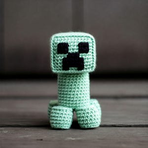 Op de afbeelding: Een groen gehaakt Minecraft Creeper knuffel met zwarte ogen en een vierkant hoofd.
