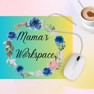 Puede incluir: Un ratón de ordenador blanco conectado a una alfombrilla con el texto "Mama's Workspace" rodeado de una corona floral de rosas azules y rosas. También se ve una taza de café con un diseño de corazón.