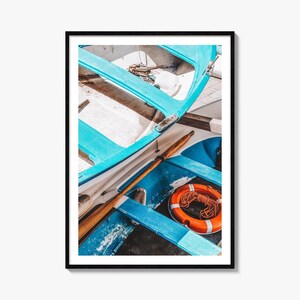 May include: A high-angle view of the interior of a bright blue wooden boat. A long wooden oar rests diagonally across the benches. An orange life ring with a coiled rope sits on the floor of the boat, which is framed by a simple black border.