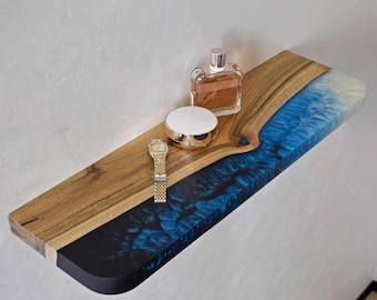 May include: A wooden floating shelf with a blue resin river design. The shelf holds a perfume bottle, a compact, and a gold wristwatch. The shelf is mounted on a white wall.