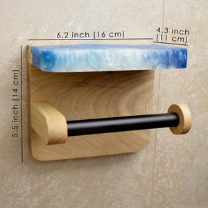 May include: A wall-mounted toilet paper holder featuring a light wood backplate, a black horizontal rod, and a top shelf made of blue and white marbled resin. The unit measures 6.2 inches wide, 4.3 inches deep, and 5.5 inches in total height.