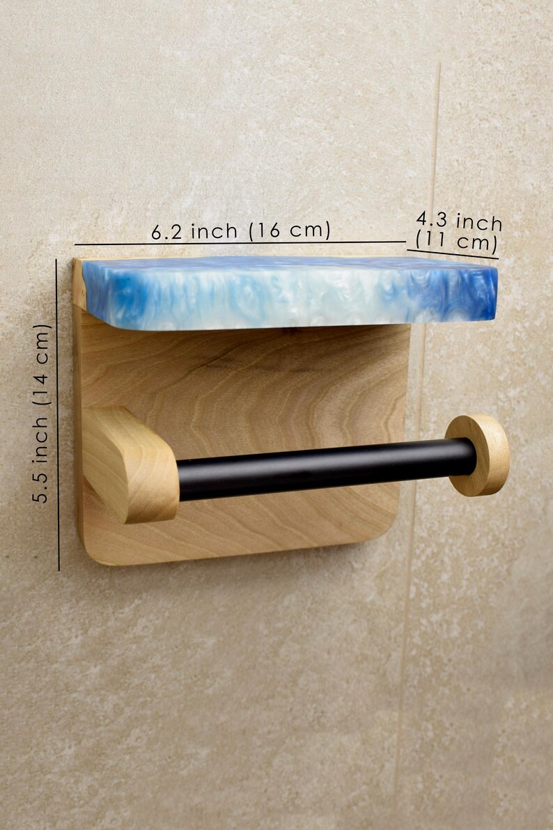 May include: A wall-mounted toilet paper holder featuring a light wood backplate, a black horizontal rod, and a top shelf made of blue and white marbled resin. The unit measures 6.2 inches wide, 4.3 inches deep, and 5.5 inches in total height.