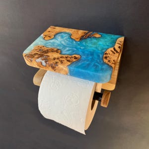 May include: A wall-mounted toilet paper holder featuring a rectangular shelf made of natural burl wood with a swirling, translucent turquoise blue resin inlay. The holder includes a wooden dowel arm and a black metal rod to support a roll of white paper.