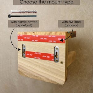 May include: A light-colored wooden wall shelf featuring two metal keyhole mounting plates on the back. The shelf includes red 3M adhesive tape strips for optional mounting and is shown with a white plastic dowel and a metal screw for installation.