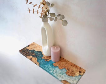 May include: A wooden shelf with a river of blue and white resin. A white vase with dried foliage and a pink candle sit on the shelf. The shelf has a natural wood grain pattern.