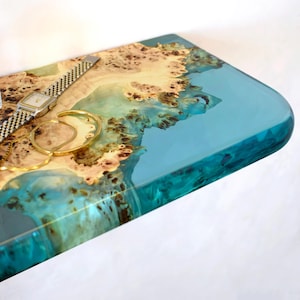 Puede incluir: Una bandeja rectangular de madera con un borde de resina transparente de color turquesa. La madera tiene un patrón natural marrón claro y beige. Un reloj plateado, una pulsera dorada y un collar dorado están en la bandeja.