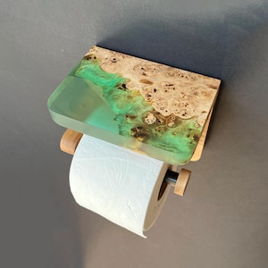 May include: A wall-mounted toilet paper holder featuring a rectangular shelf made of natural wood burl and translucent green resin. The holder includes a wooden dowel arm that supports a roll of white toilet paper with a subtle embossed floral pattern.