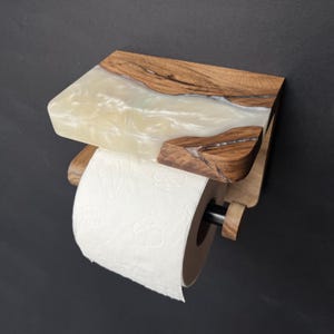 May include: A wall-mounted toilet paper holder featuring a natural wood shelf with a pearlescent white resin inlay. The holder includes a black metal rod supporting a roll of white toilet paper with a subtle embossed floral pattern on the surface.