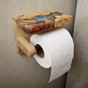May include: A wall-mounted toilet paper holder featuring a light-colored wooden base and a shelf top. The shelf is constructed from natural wood burl with a central inlay of blue and reddish-brown resin. A black rod holds a roll of white toilet paper.