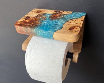 May include: A wall-mounted toilet paper holder featuring a natural wood shelf with a vibrant blue resin inlay that mimics a flowing river. The holder includes a horizontal wooden rod with a black accent, supporting a roll of white embossed toilet paper.