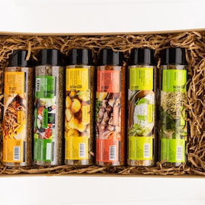 May include: A gift set of six spice jars with black lids, each with a colorful label and a picture of a dish. The jars are arranged in a brown cardboard box filled with shredded paper.