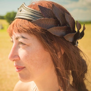 May include: A woman with auburn hair wearing a silver-toned crown and a dark brown leaf-shaped headpiece. The headpiece is made of leather and is in the shape of a laurel wreath. The background is a sunny field.