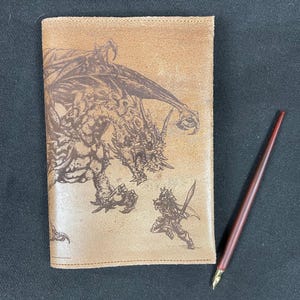 May include: A brown leather journal with a dragon and a knight design. The journal is closed and has a pen next to it.