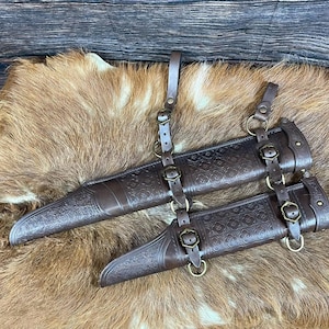 Puede incluir: Un conjunto de fundas de cuero marrón oscuro con intrincados patrones florales en relieve. Las fundas están conectadas por correas de cuero ajustables con hebillas y anillos de metal en tono latón antiguo para su sujeción.