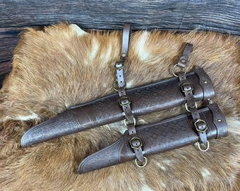 May include: A dark brown leather scabbard set featuring intricate embossed floral patterns. The scabbards are connected by adjustable leather straps with antique brass-toned metal buckles and rings, designed for secure attachment and carrying.