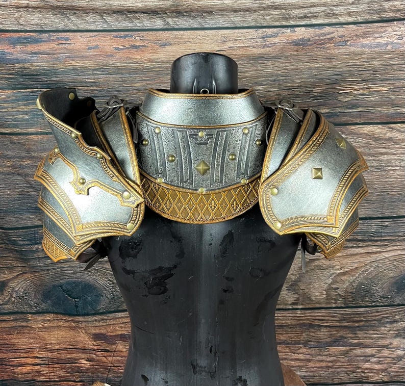 May include: A silver and gold-colored shoulder armor set. The armor features intricate detailing, including rivets, decorative patterns, and layered plates. The design suggests a medieval or fantasy theme, with a weathered appearance.