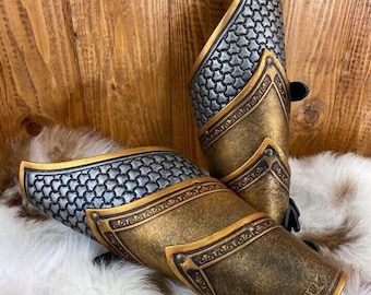 May include: A pair of gold and silver forearm armor with chainmail detailing. The armor is made of leather and has a distressed finish.