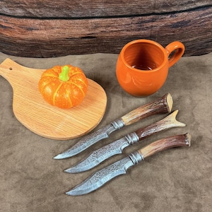 Peut inclure: Trois couteaux avec des lames en métal texturé et des manches en bois façon bois de cerf disposés en diagonale. À côté se trouvent une petite tasse en céramique orange et une citrouille décorative orange sur une planche à découper en bois.