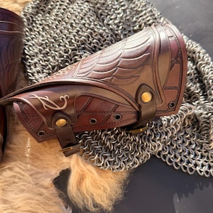 Puede incluir: Primer plano de un protector de brazo de cuero marr&oacute;n con dise&ntilde;o de alas en relieve y detalles decorativos. El protector de brazo tiene un cierre de hebilla y est&aacute; colocado sobre una cota de malla y un fondo texturizado. Otro protector de brazo es parcialmente visible.