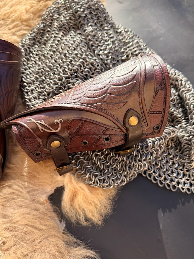 Puede incluir: Primer plano de un protector de brazo de cuero marr&oacute;n con dise&ntilde;o de alas en relieve y detalles decorativos. El protector de brazo tiene un cierre de hebilla y est&aacute; colocado sobre una cota de malla y un fondo texturizado. Otro protector de brazo es parcialmente visible.