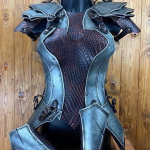 Pode incluir: Um conjunto de armadura de couro azul e marrom com um padrão de crocodilo. A armadura inclui uma peça de peito, ombreiras e protetores de braço.