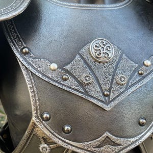May include: Close-up of a black and silver chest armor. The armor features intricate floral and geometric patterns, with a central decorative emblem. Rivets and detailed edging add to the medieval aesthetic.