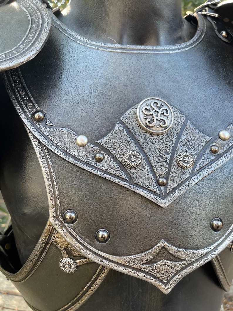 May include: Close-up of a black and silver chest armor. The armor features intricate floral and geometric patterns, with a central decorative emblem. Rivets and detailed edging add to the medieval aesthetic.