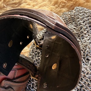 Puede incluir: Primer plano de una gola de cuero marr&oacute;n con remaches de lat&oacute;n y un interior negro. La gola forma parte de un conjunto de armadura medieval, que descansa sobre una superficie de cota de malla. El cuero tiene un aspecto texturizado.