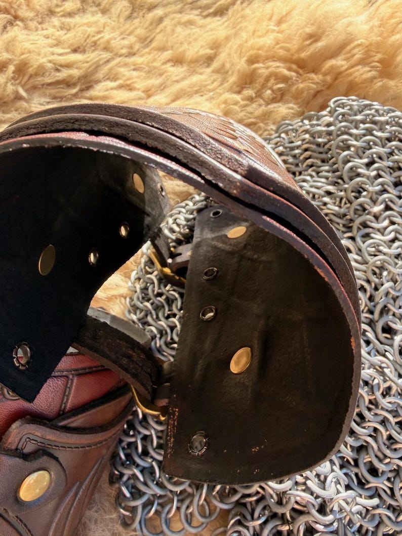 Puede incluir: Primer plano de una gola de cuero marr&oacute;n con remaches de lat&oacute;n y un interior negro. La gola forma parte de un conjunto de armadura medieval, que descansa sobre una superficie de cota de malla. El cuero tiene un aspecto texturizado.