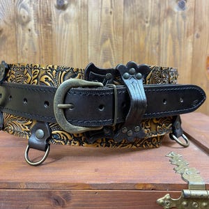 May include: A brown leather belt with a gold-toned embossed design and a metal buckle. The belt has two D-rings on either side.
