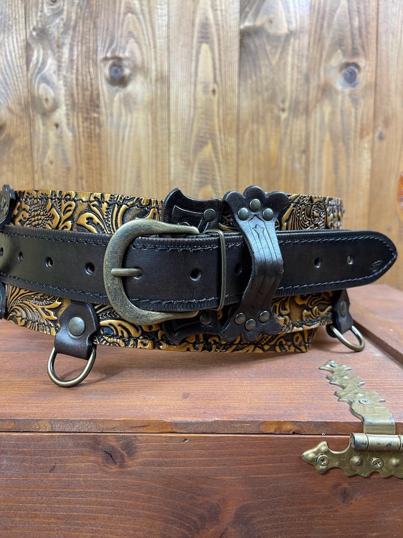 May include: A brown leather belt with a gold-toned embossed design and a metal buckle. The belt has two D-rings on either side.