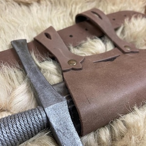 Könnte beinhalten: Ein braunes Lederschwert-Scheide mit einem Metallsäbel im Inneren. Die Scheide hat eine Gürtelschlaufe und zwei Riemen. Das Schwert hat einen seilumwickelten Griff.