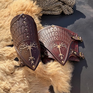 Puede incluir: Un par de protectores de brazo de cuero marr&oacute;n con intrincados dise&ntilde;os en relieve. La armadura presenta un emblema del &aacute;rbol de la vida y patrones de estrellas. El cuero es de un marr&oacute;n intenso con detalles dorados y hebillas de lat&oacute;n. El fondo incluye un material similar a la piel y cota de malla.