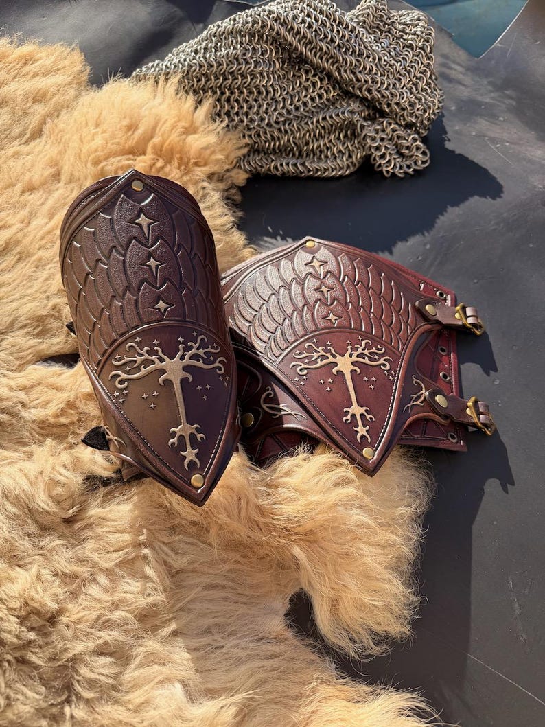 Puede incluir: Un par de protectores de brazo de cuero marr&oacute;n con intrincados dise&ntilde;os en relieve. La armadura presenta un emblema del &aacute;rbol de la vida y patrones de estrellas. El cuero es de un marr&oacute;n intenso con detalles dorados y hebillas de lat&oacute;n. El fondo incluye un material similar a la piel y cota de malla.