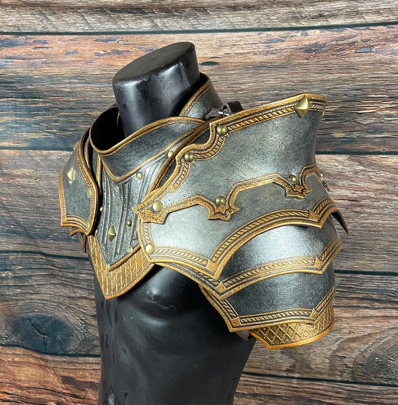 May include: A detailed, metallic shoulder armor piece in silver and gold tones. The armor features intricate embossed designs, rivets, and decorative edges. The piece is displayed on a black mannequin, showcasing its form and craftsmanship. The armor is designed for cosplay or costume use.