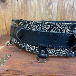 May include: A black leather belt with a silver embossed floral pattern. The belt has two metal D-rings and a buckle with a decorative design.