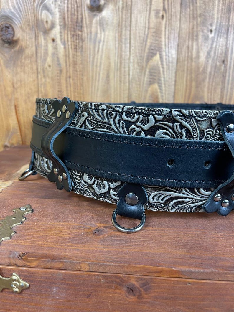 May include: A black leather belt with a silver embossed floral pattern. The belt has two metal D-rings and a buckle with a decorative design.