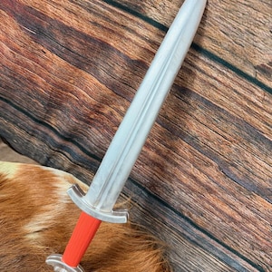 May include: A toy sword with a silver blade and a red handle. The sword has a detailed hilt and a pointed tip. The toy is made of plastic and is designed to look like a Viking sword.