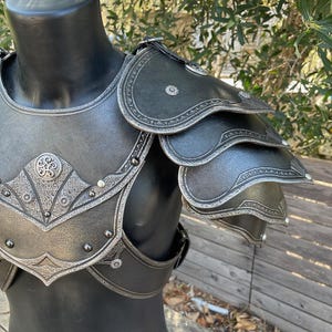 May include: A black and silver shoulder armor set displayed on a mannequin. The armor features intricate embossed designs and rivets, with layered shoulder pauldrons. The overall aesthetic suggests a medieval or fantasy theme.