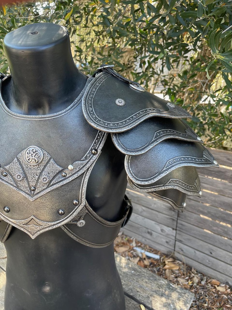 May include: A black and silver shoulder armor set displayed on a mannequin. The armor features intricate embossed designs and rivets, with layered shoulder pauldrons. The overall aesthetic suggests a medieval or fantasy theme.