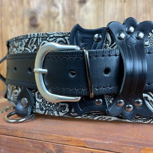 May include: A black leather belt with a silver buckle and decorative metal accents. The belt has a floral pattern embossed on the leather.