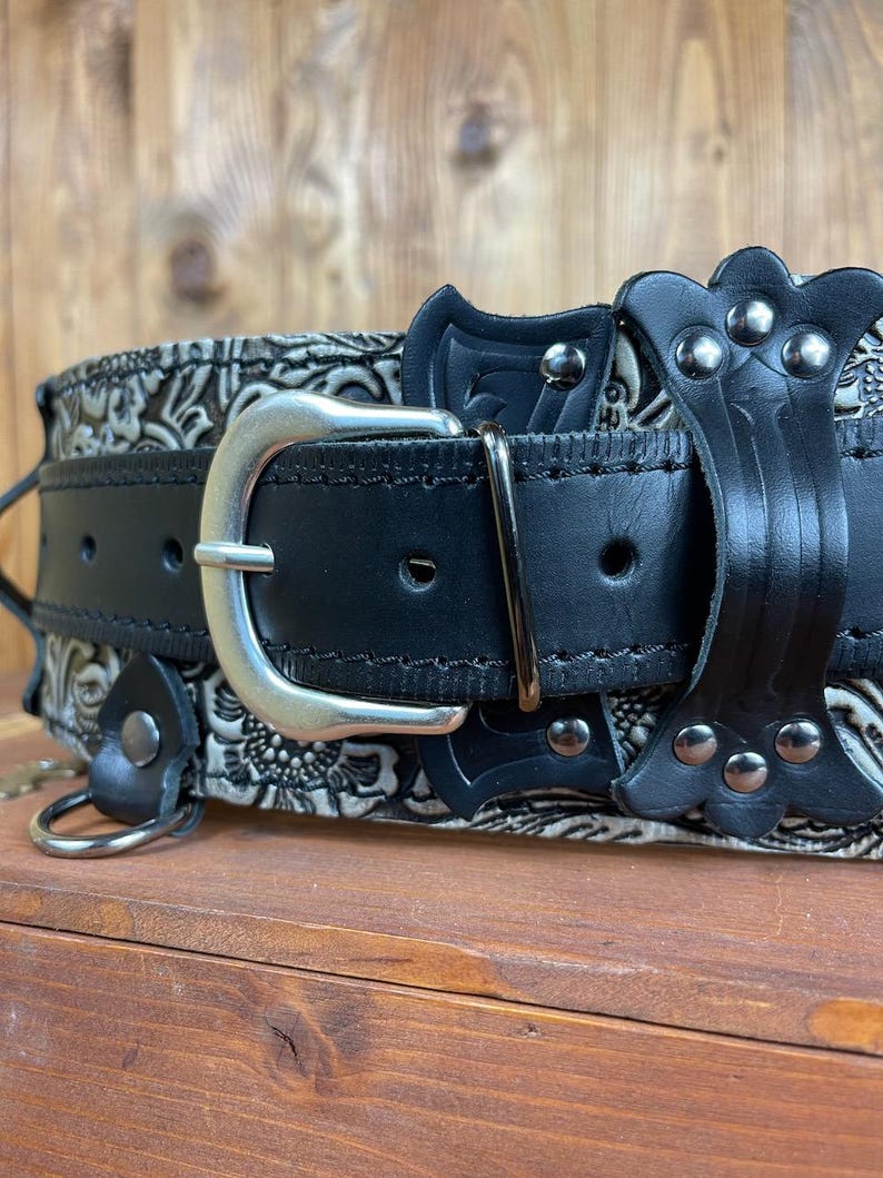 May include: A black leather belt with a silver buckle and decorative metal accents. The belt has a floral pattern embossed on the leather.