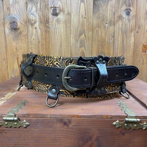 May include: A black leather belt with a gold embossed floral design. The belt has a buckle and D-rings.