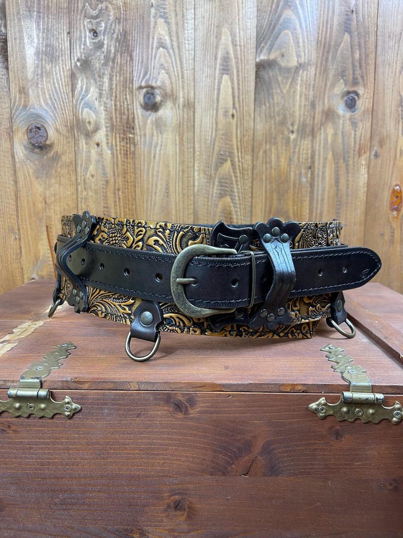May include: A black leather belt with a gold embossed floral design. The belt has a buckle and D-rings.