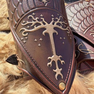 Puede incluir: Primer plano de protectores de brazo de cuero marr&oacute;n con patrones de escamas y estrellas en relieve. El dise&ntilde;o central presenta un &aacute;rbol dorado con estrellas. Un tachuela dorada est&aacute; en la parte inferior. Los protectores de brazo son probablemente para cosplay o recreaciones hist&oacute;ricas.