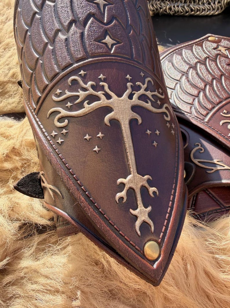 Puede incluir: Primer plano de protectores de brazo de cuero marr&oacute;n con patrones de escamas y estrellas en relieve. El dise&ntilde;o central presenta un &aacute;rbol dorado con estrellas. Un tachuela dorada est&aacute; en la parte inferior. Los protectores de brazo son probablemente para cosplay o recreaciones hist&oacute;ricas.