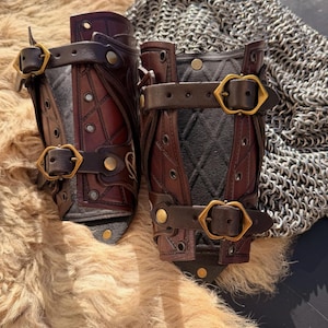 Puede incluir: Par de protectores de brazo de cuero hechos a mano en tonos marr&oacute;n y negro, con costuras intrincadas y hebillas de lat&oacute;n. Los protectores de brazo tienen un interior acolchado. El fondo incluye una cota de malla y una superficie peluda.
