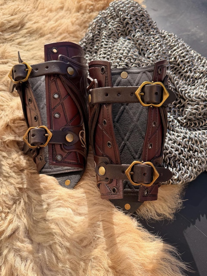 Puede incluir: Par de protectores de brazo de cuero hechos a mano en tonos marr&oacute;n y negro, con costuras intrincadas y hebillas de lat&oacute;n. Los protectores de brazo tienen un interior acolchado. El fondo incluye una cota de malla y una superficie peluda.