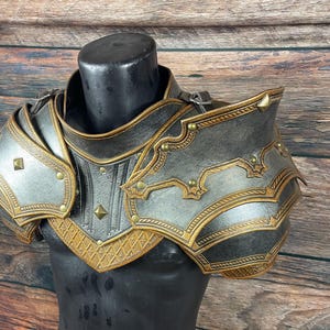 May include: A detailed, handcrafted shoulder armor set in shades of gray and gold, displayed on a black mannequin. The armor features intricate embossed designs, rivets, and decorative edging, showcasing a medieval or fantasy theme.