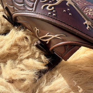 Puede incluir: Primer plano de protectores de brazo de cuero marr&oacute;n con dise&ntilde;os en relieve. Los protectores de brazo presentan patrones intrincados y un motivo de &aacute;rbol y estrellas de color dorado. El cuero tiene un color marr&oacute;n oscuro intenso con acentos marrones claros.